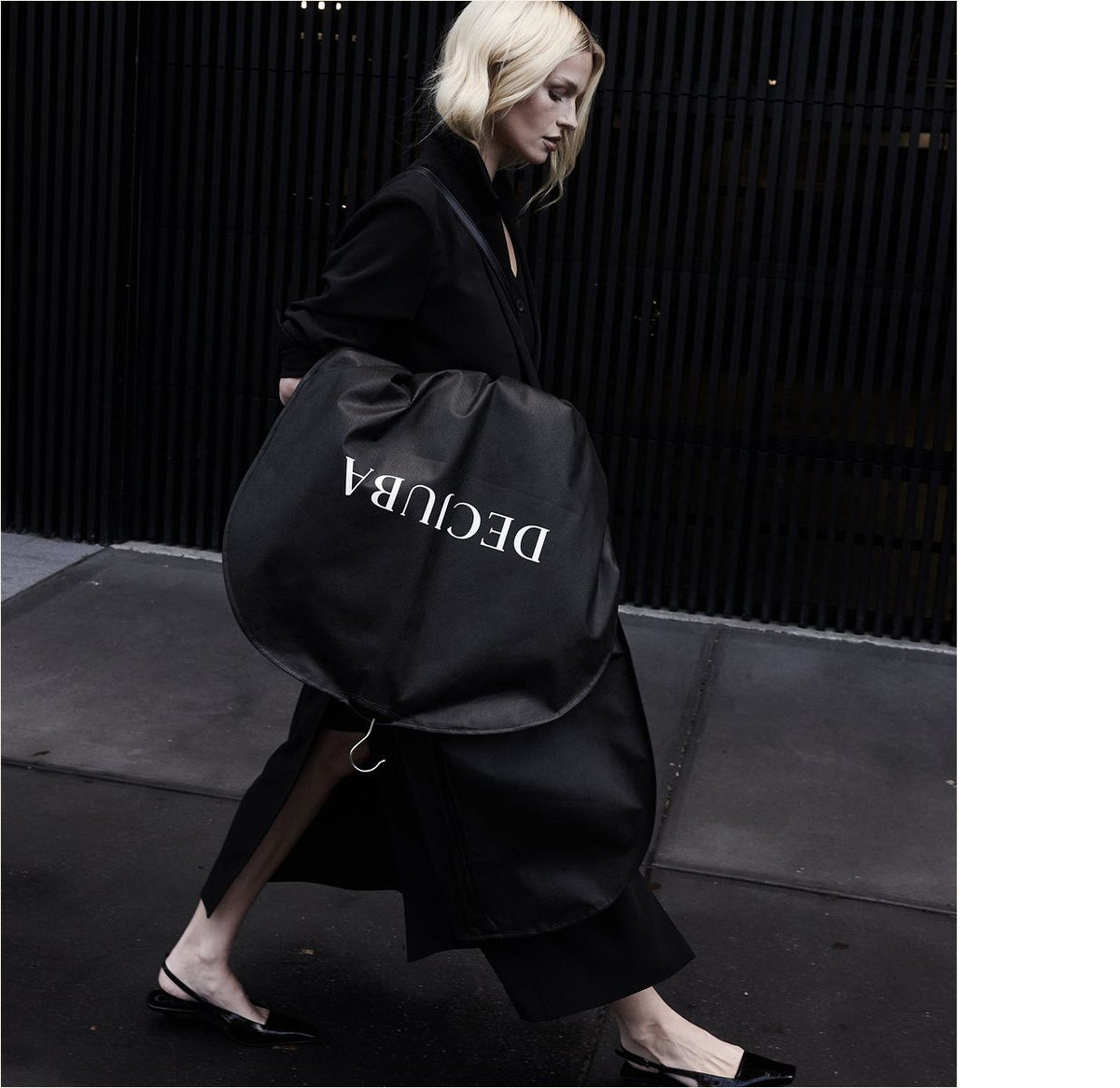 A woman in a sleek black outfit walks outside with a large, round black coat bag labelled "DECUBA"