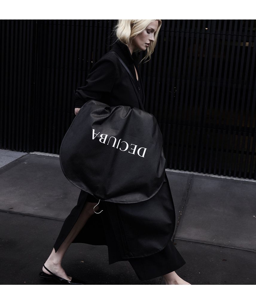 A woman in a sleek black outfit walks outside with a large, round black coat bag labelled "DECUBA"